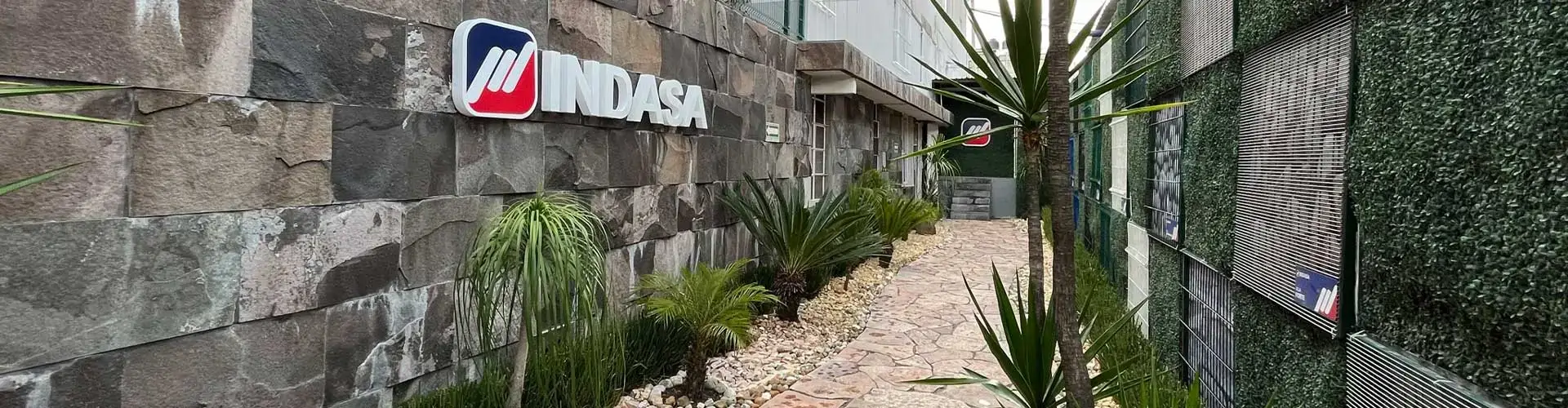 Pasillo exterior con paredes de piedra y plantas verdes decorativas, con el logotipo de INDASA montado en una pared.