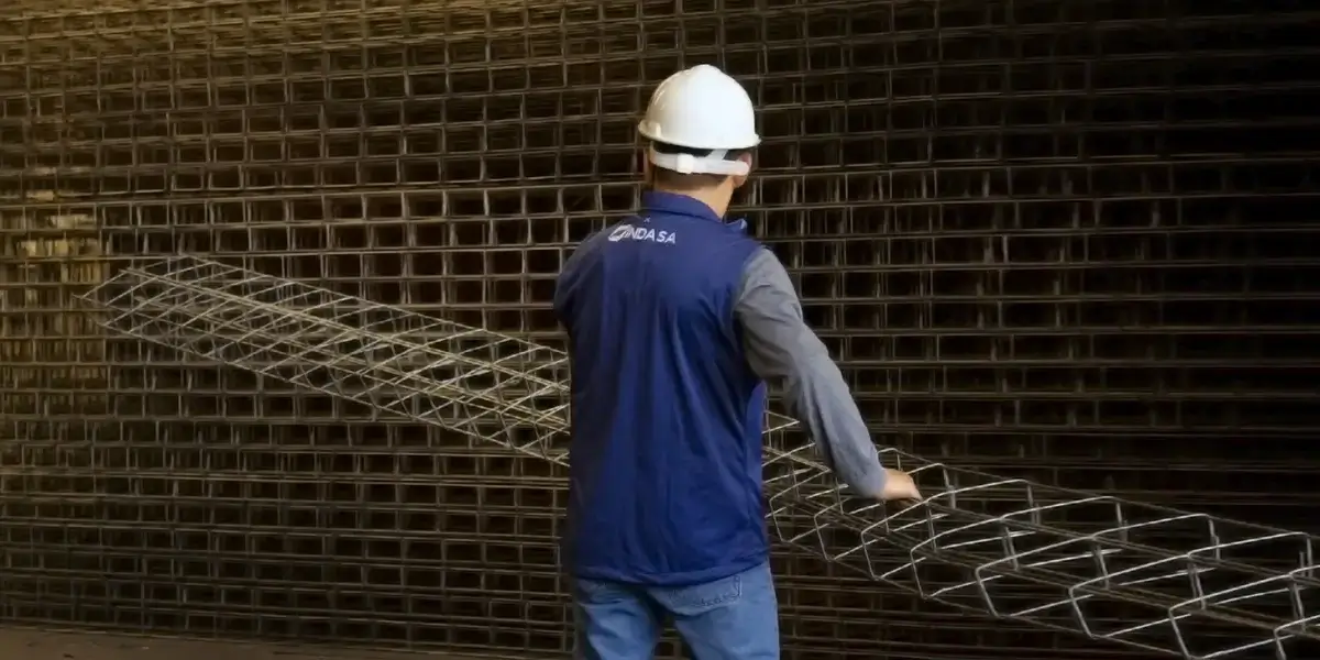 Trabajador con casco blanco y chaleco azul de INDASA cargando una castillos metálico frente a un fondo de castillos apiladas